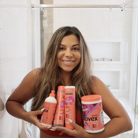 Evelyn Regly está no banheiro segurando toda a família Novex Infusão de Colágeno composta por: shampoo, creme de tratamento, tratamento condicionante e creme para pentear.