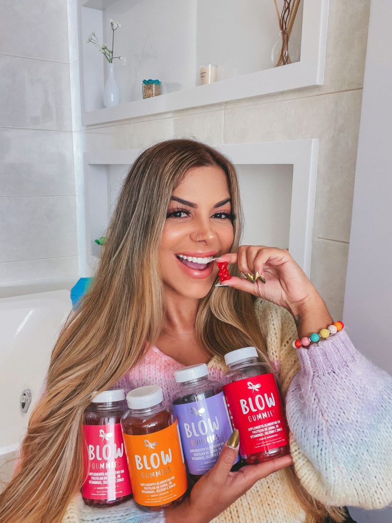 Evelyn Regly está usando um casaco colorido e segura no colo quatro frascos de gominhas Blow Gummies. Uma de cada sabor: morango, laranja, uva e melancia. Em uma das mãos segura próximo ao rosto uma gominha do sabor melancia. A gominha tem formato de ursinho.